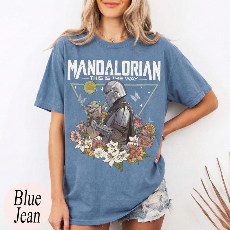 May include: Blue jean t-shirt featuring a graphic of The Mandalorian and Grogu surrounded by flowers and butterflies. The text "MANDALORIAN - THIS IS THE WAY" is printed across the top. The shirt is a light blue color.