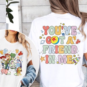 May include: White t-shirt featuring a colorful graphic of Toy Story characters, including Woody, Buzz Lightyear, and Rex. The back of the shirt displays the text "YOU'VE GOT A FRIEND IN ME" in a retro font with stars and space-themed illustrations.