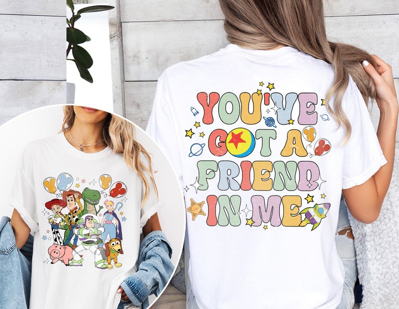 May include: White t-shirt featuring a colorful graphic of Toy Story characters, including Woody, Buzz Lightyear, and Rex. The back of the shirt displays the text "YOU'VE GOT A FRIEND IN ME" in a retro font with stars and space-themed illustrations.