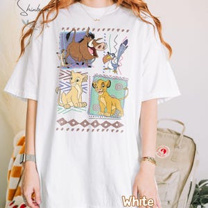 May include: White t-shirt featuring a colorful graphic of characters from The Lion King. The design includes images of Simba, Timon, Pumbaa, and Zazu, set within decorative frames. The word "White" is printed at the bottom.