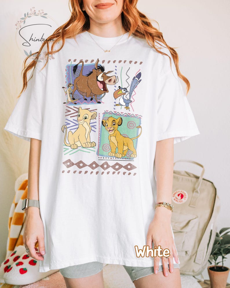 May include: White t-shirt featuring a colorful graphic of characters from The Lion King. The design includes images of Simba, Timon, Pumbaa, and Zazu, set within decorative frames. The word "White" is printed at the bottom.