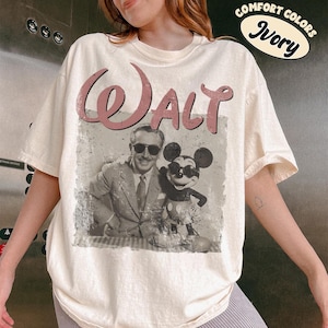 May include: Ivory-colored t-shirt featuring a vintage-style graphic of Walt Disney and Mickey Mouse. The word "Walt" is printed in a dusty rose color above the image. The shirt has a relaxed fit and short sleeves.