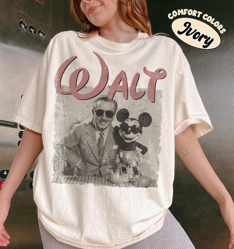 May include: Ivory-colored t-shirt featuring a vintage-style graphic of Walt Disney and Mickey Mouse. The word "Walt" is printed in a dusty rose color above the image. The shirt has a relaxed fit and short sleeves.