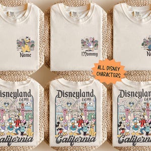 May include: Cream-colored t-shirts with Disney character graphics. Some shirts have "Name" or "Mommy" text. The bottom shirts feature a Disneyland graphic with "California" text and "Est. 1955". The shirts are displayed on woven mats.