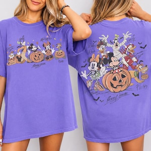 May include: Purple t-shirt featuring a Halloween-themed graphic with Mickey Mouse, Minnie Mouse, Goofy, Donald Duck, and Pluto characters. The front and back of the shirt display the characters with pumpkins and bats.