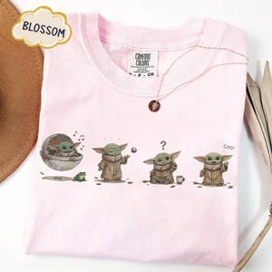 Puede incluir: Camiseta rosa claro con un gr&aacute;fico de un personaje de una popular serie de ciencia ficci&oacute;n. El gr&aacute;fico muestra al personaje en diferentes poses. La camiseta tambi&eacute;n tiene la palabra "BLOSSOM" en un dise&ntilde;o floral.