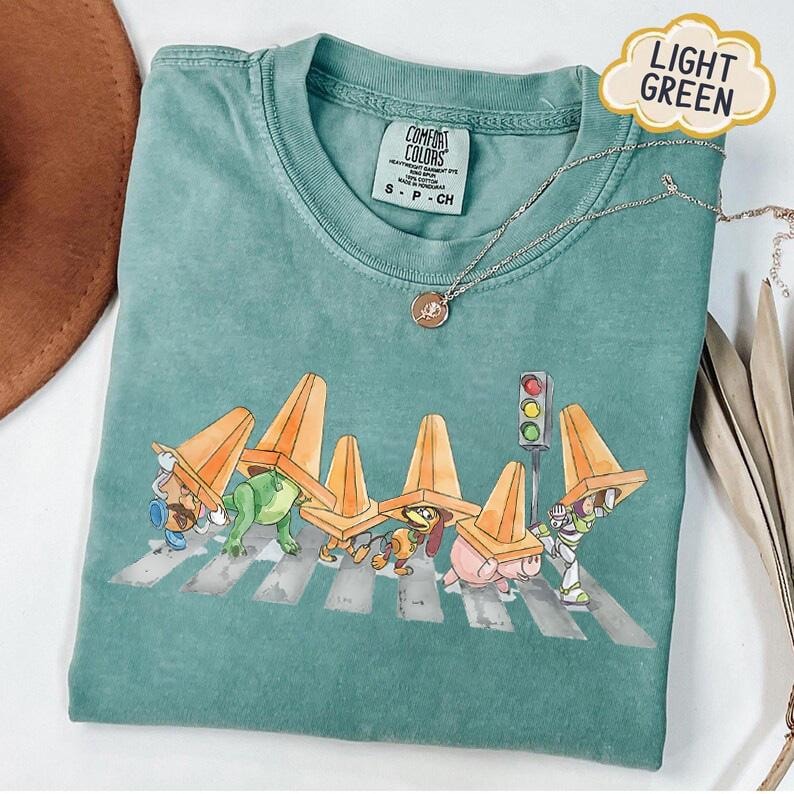 May include: Light green Comfort Colors t-shirt featuring a cartoon graphic of characters crossing a crosswalk, wearing orange traffic cones. The shirt has a crew neck and short sleeves. A silver necklace with a pendant is also visible.
