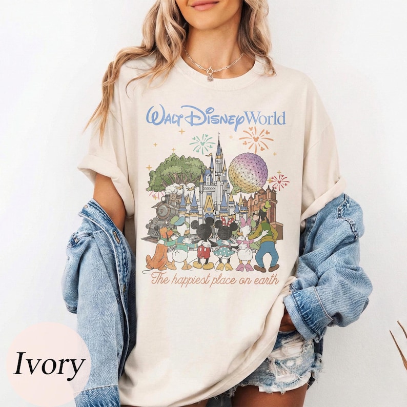 Puede incluir: Camiseta de color marfil con un gr&aacute;fico colorido de Walt Disney World con personajes y lugares emblem&aacute;ticos. El texto "Walt Disney World" est&aacute; encima del gr&aacute;fico y "The happiest place on earth" debajo. La camiseta se lleva con una chaqueta vaquera.