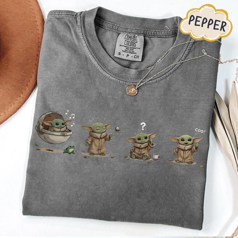 Puede incluir: Camiseta gris Comfort Colors con un gr&aacute;fico de un personaje con una capa marr&oacute;n en cuatro poses diferentes. El personaje se muestra con una rana, un signo de interrogaci&oacute;n y una taza de caf&eacute;. Tambi&eacute;n se ven un collar plateado y una etiqueta Pepper.