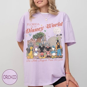 May include: A lilac t-shirt with a vibrant graphic of Disney World characters and attractions. The design showcases Mickey Mouse, Minnie Mouse, Donald Duck, and Goofy, with the text "Florida Disney World" and "The Most Magical Place On Earth."