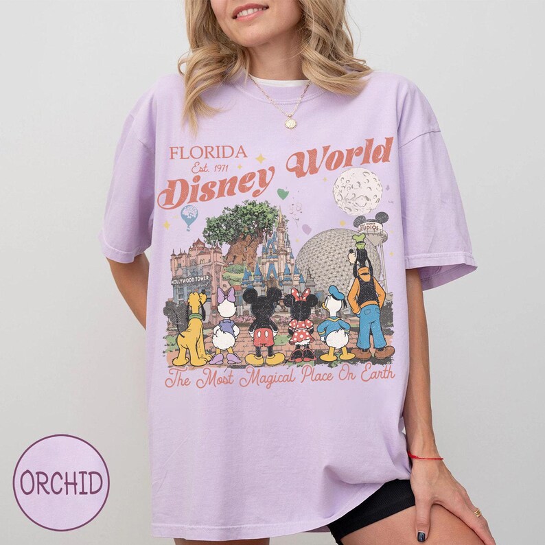 May include: A lilac t-shirt with a vibrant graphic of Disney World characters and attractions. The design showcases Mickey Mouse, Minnie Mouse, Donald Duck, and Goofy, with the text "Florida Disney World" and "The Most Magical Place On Earth."