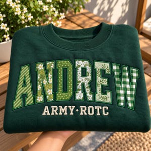 Könnte beinhalten: Ein dunkelgrüner Rundhals-Pullover mit dem Namen ANDREW in großen Blockbuchstaben. Jeder Buchstabe hat ein einzigartiges Muster in Grüntönen, darunter Streifen, Gänseblümchen, Punkte, Paisley, Blätter und Vichy-Karo. Darunter ist ARMY ROTC eingestickt.