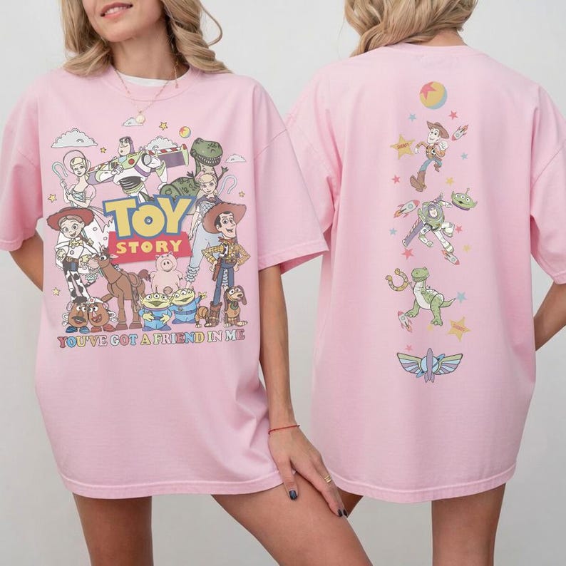 Op de afbeelding: Een lichtroze T-shirt met een Toy Story-afbeelding op de voor- en achterkant. De voorkant toont personages zoals Woody, Buzz Lightyear en Jessie met de tekst Toy Story en You've Got A Friend In Me. De achterkant toont een verticale opstelling van personages en sterren.