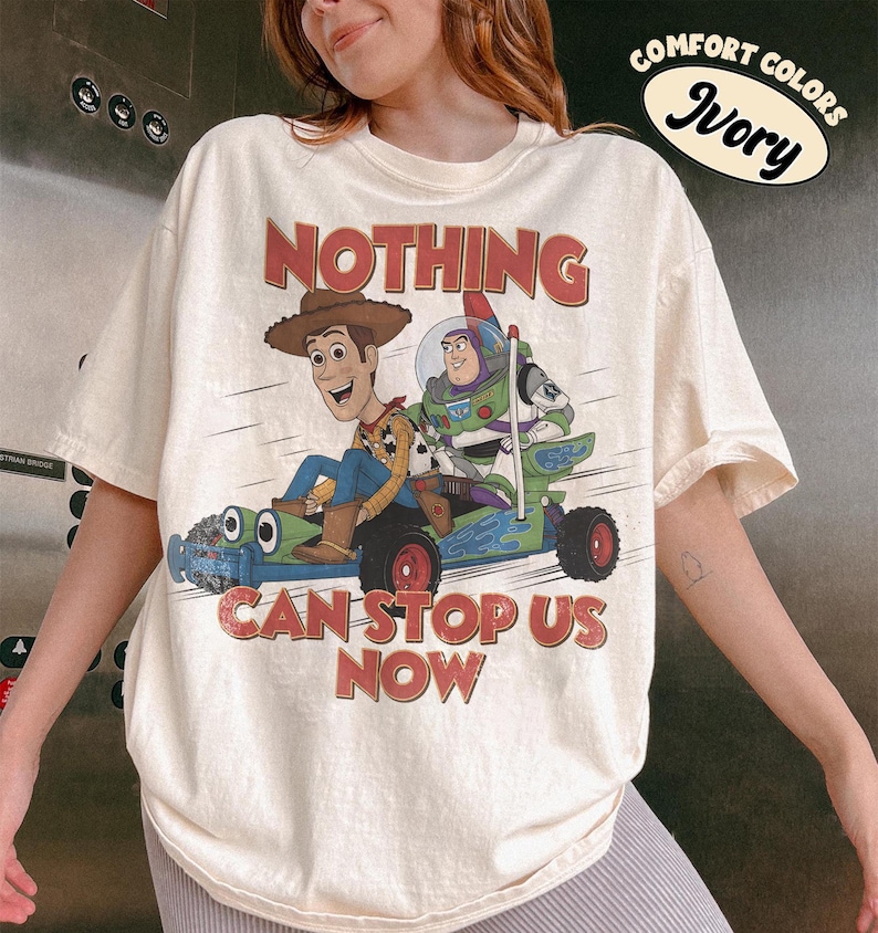 May include: An ivory short-sleeve t-shirt featuring a graphic of Woody and Buzz Lightyear riding a green toy car. The text reads "NOTHING CAN STOP US NOW" in red letters. A "COMFORT COLORS IVORY" label is visible in the upper right corner.