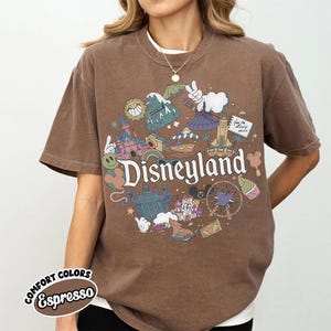 May include: Espresso-colored t-shirt featuring a circular Disneyland graphic with iconic park imagery. The design includes a castle, sun, and other park elements. The shirt has short sleeves and a crew neck.