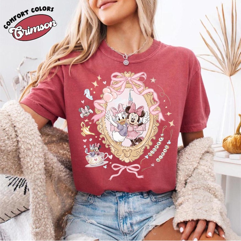 May include: Crimson Comfort Colors t-shirt featuring a framed illustration of Daisy Duck and Minnie Mouse. The design includes bows, stars, and other Disney-themed elements. The shirt has short sleeves and a crew neck.