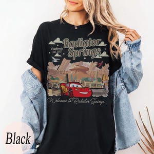 May include: Black t-shirt with a graphic of Radiator Springs, including the text "Radiator Springs" and "Welcome to Radiator Springs." The design features a red race car and a desert scene. The shirt also has the text "California est. 2012."