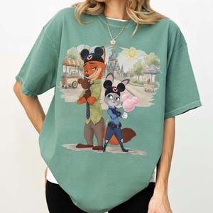 Puede incluir: Camiseta verde con una ilustraci&oacute;n de dibujos animados de Nick Wilde y Judy Hopps de Zootopia. Los personajes est&aacute;n frente a una escena de parque con un castillo y algod&oacute;n de az&uacute;car. La camiseta tiene un corte holgado.