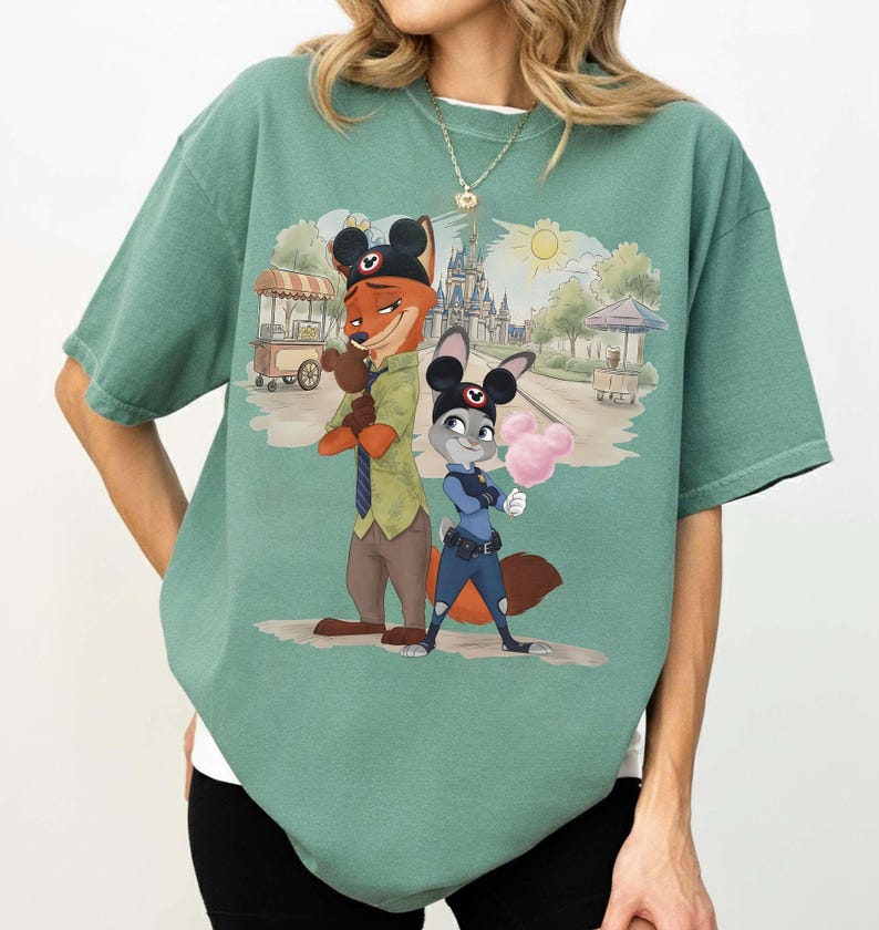 Puede incluir: Camiseta verde con una ilustraci&oacute;n de dibujos animados de Nick Wilde y Judy Hopps de Zootopia. Los personajes est&aacute;n frente a una escena de parque con un castillo y algod&oacute;n de az&uacute;car. La camiseta tiene un corte holgado.