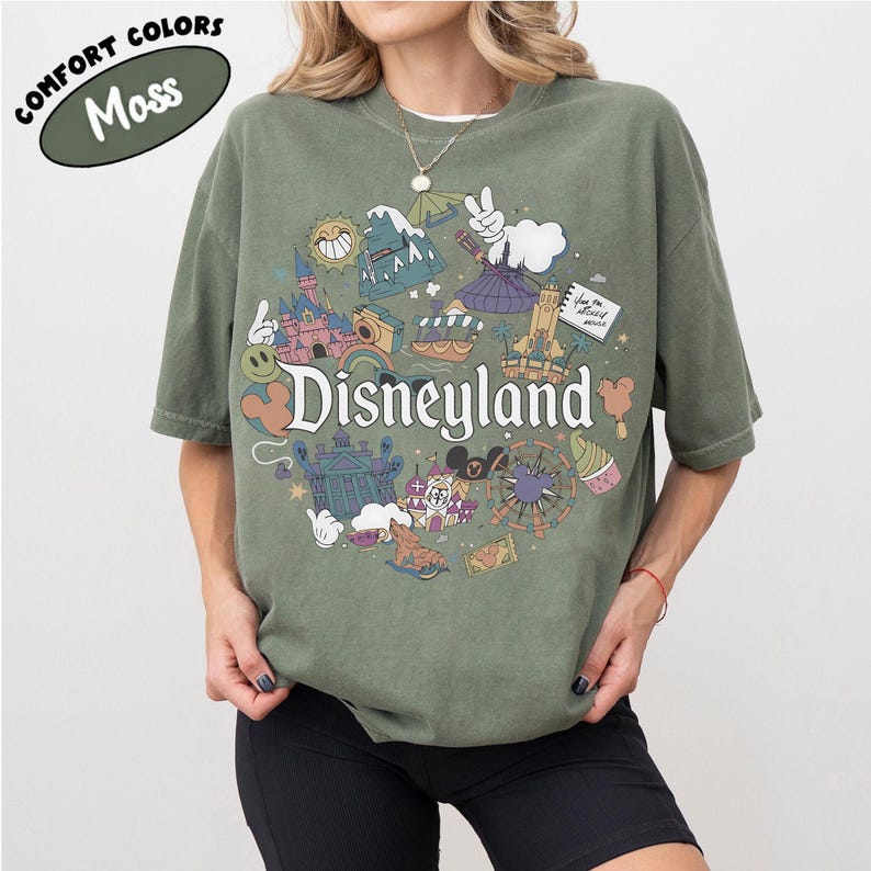 May include: A moss green t-shirt with a Disneyland graphic. The graphic features various Disneyland icons, including the castle, a smiling sun, and a Ferris wheel. The words "Disneyland" are prominently displayed.