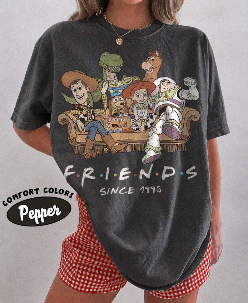 May include: A charcoal gray short-sleeve t-shirt featuring a graphic of Toy Story characters sitting on a couch. The text reads "F R I E N D S" and "SINCE 1995" in white. A small oval logo on the side displays the text "COMFORT COLORS" and "Pepper".