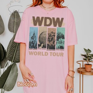 May include: Pink t-shirt with a vintage-style graphic of the four theme parks at Walt Disney World. The graphic includes the park names: Magic Kingdom, Epcot Center, Hollywood Studios, and Animal Kingdom. The text "WDW World Tour" is printed below the graphic.