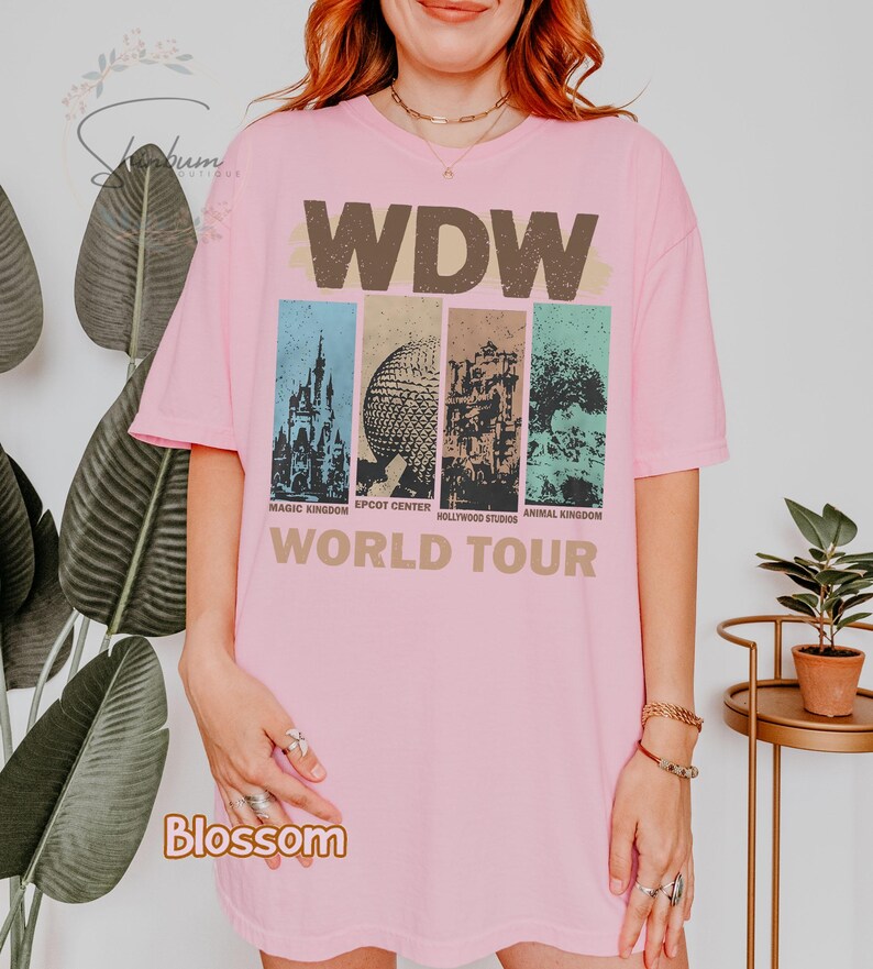 May include: Pink t-shirt with a vintage-style graphic of the four theme parks at Walt Disney World. The graphic includes the park names: Magic Kingdom, Epcot Center, Hollywood Studios, and Animal Kingdom. The text "WDW World Tour" is printed below the graphic.