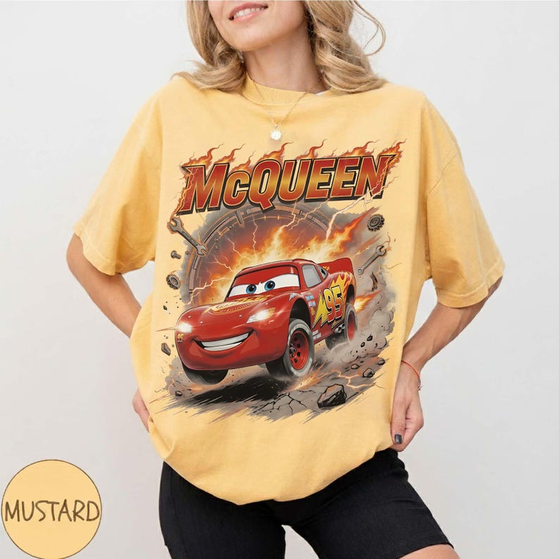 Może przedstawiać: Musztardowa koszulka z grafiką Zygzaka McQueena z filmu Auta. Projekt zawiera napis "McQueen" w płomieniach nad czerwonym samochodem wyścigowym. Koszulka ma luźny, obszerny kr&oacute;j.