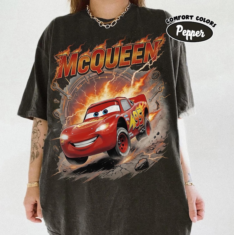 Może przedstawiać: Czarny t-shirt z grafiką Zygzaka McQueena z filmu Auta. Projekt zawiera słowo "McQueen" w kolorze pomarańczowym, samoch&oacute;d wyścigowy i numer 95. Koszulka ma r&oacute;wnież napis "Comfort Colors Pepper".