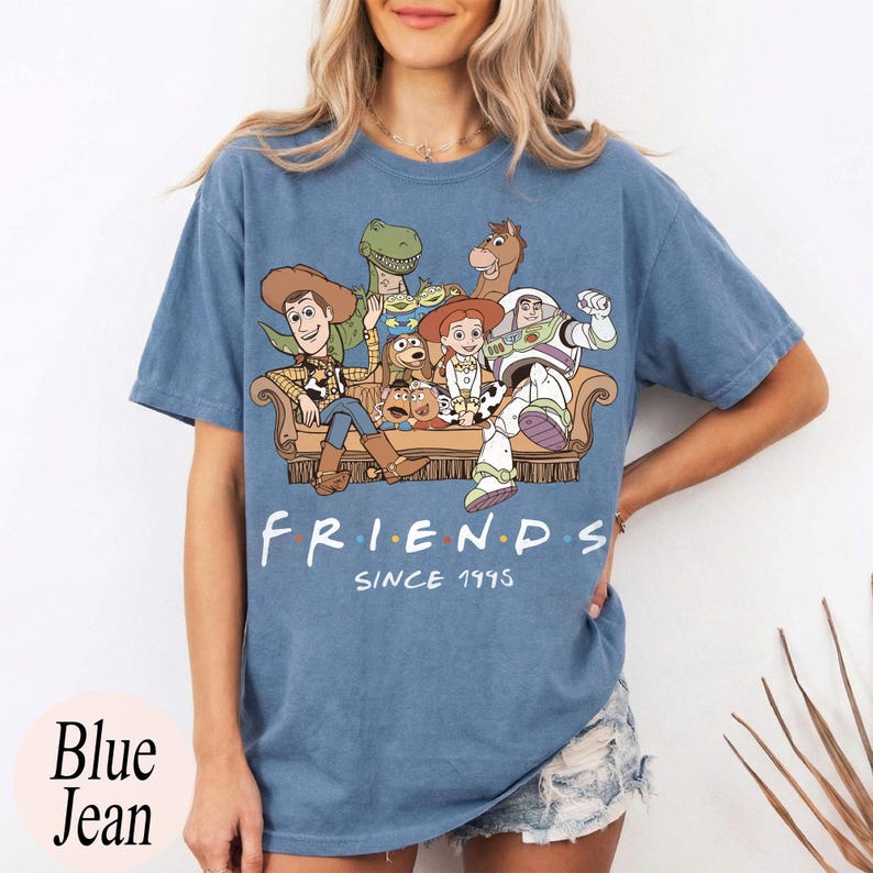 May include: A blue short-sleeve t-shirt featuring a graphic print of Toy Story characters sitting on a couch. The text below the characters reads "FRIENDS" with colored dots between letters and "SINCE 1995" written in a white script font below.