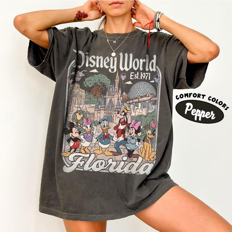 May include: Dark gray t-shirt featuring a Disney World graphic with Mickey Mouse, Minnie Mouse, Donald Duck, Daisy Duck, Goofy, and Pluto. The graphic includes the text "Disney World Est. 1971 Florida". The shirt is labeled "Comfort Colors Pepper".