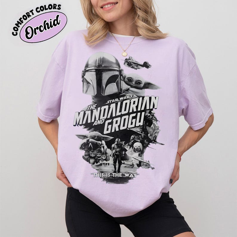 Op de afbeelding: Een lichtpaars T-shirt met een afbeelding uit de Star Wars-serie, "The Mandalorian and Grogu". Het ontwerp bevat afbeeldingen van de Mandalorian, Grogu en ruimtevaartuigen. De tekst "THIS IS THE WAY" is ook aanwezig.