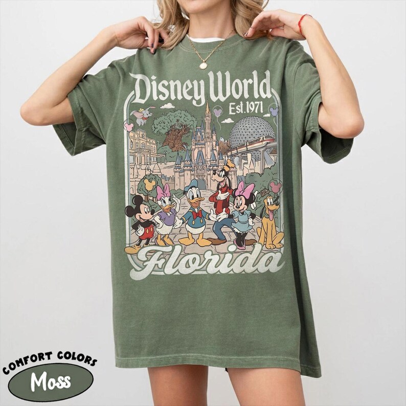 May include: Moss green t-shirt featuring a graphic of Disney World in Florida. The design includes Mickey Mouse, Minnie Mouse, Donald Duck, Goofy, and other characters. The text "Disney World Est. 1971" and "Florida" are also visible.