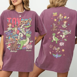 May include: A maroon-colored t-shirt featuring the words "Toy Story" in a retro font, along with illustrations of characters like Woody, Buzz, and Rex. The back of the shirt displays additional character graphics and the "Toy Story" logo.