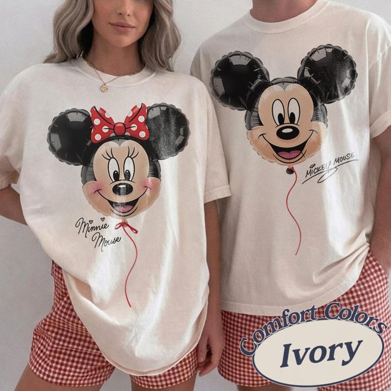 K&ouml;nnte beinhalten: Elfenbeinfarbene T-Shirts mit Mickey- und Minnie-Mouse-Ballon-Designs. Minnie's Ballon hat eine rote Schleife, Mickey's ist schlicht. Auf den Shirts stehen die Worte "Minnie Mouse" und "Mickey Mouse".