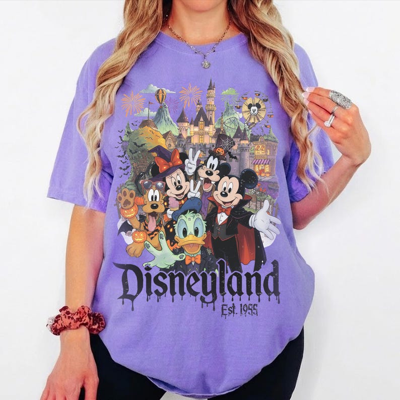 May include: A light purple short-sleeve t-shirt featuring a graphic of Disney characters in Halloween costumes in front of a castle. The text reads Disneyland Est. 1955. The design includes Mickey, Minnie, Donald, and Pluto with pumpkins.