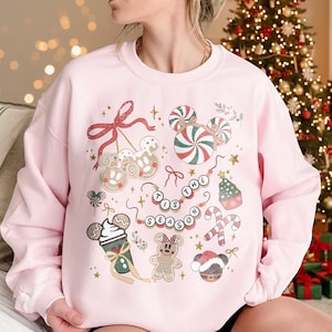 May include: A light pink sweatshirt with a festive holiday design. The design includes candy canes, gingerbread characters, and the phrase "'Tis the Season" in a circular pattern. Gold stars and bows complete the holiday theme.