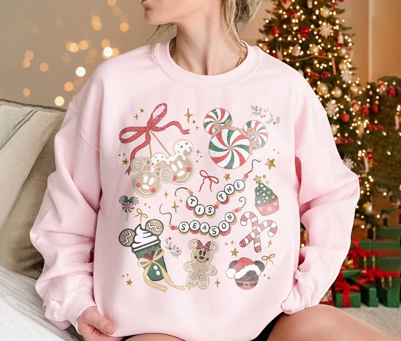 May include: A light pink sweatshirt with a festive holiday design. The design includes candy canes, gingerbread characters, and the phrase "'Tis the Season" in a circular pattern. Gold stars and bows complete the holiday theme.