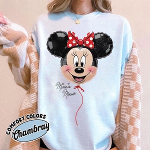 K&ouml;nnte beinhalten: Hellblaues Comfort Colors Chambray Sweatshirt mit einem Minnie Mouse Ballon-Design. Der Ballon hat schwarze Ohren, eine rote Schleife mit wei&szlig;en Punkten und ein l&auml;chelndes Gesicht. Die Worte "Minnie Mouse" sind kursiv geschrieben.