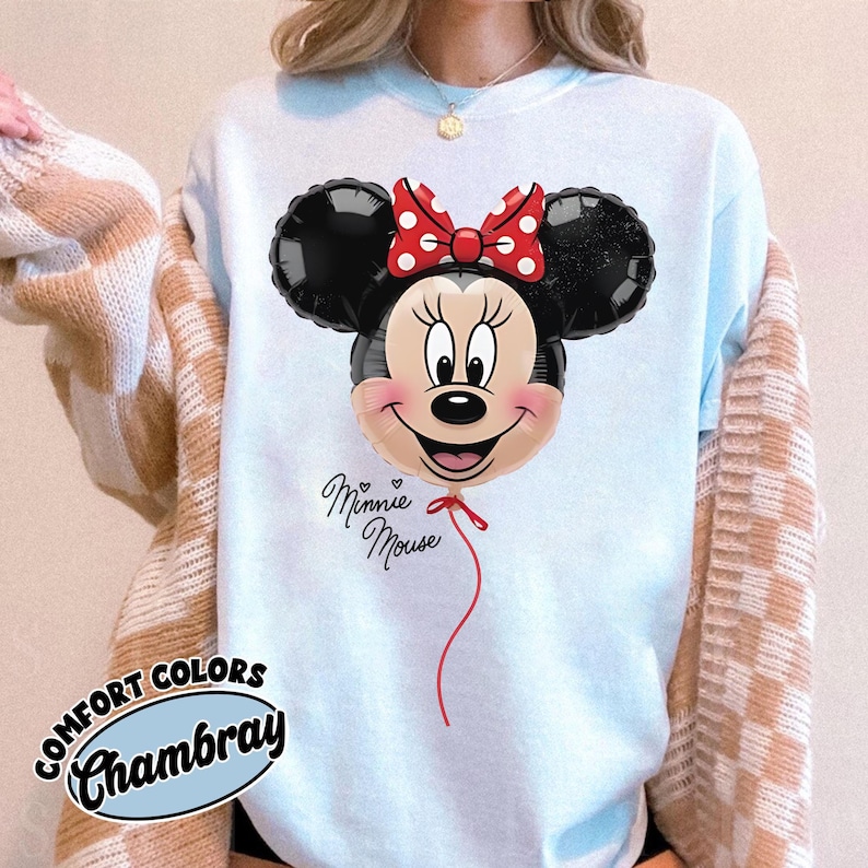 K&ouml;nnte beinhalten: Hellblaues Comfort Colors Chambray Sweatshirt mit einem Minnie Mouse Ballon-Design. Der Ballon hat schwarze Ohren, eine rote Schleife mit wei&szlig;en Punkten und ein l&auml;chelndes Gesicht. Die Worte "Minnie Mouse" sind kursiv geschrieben.