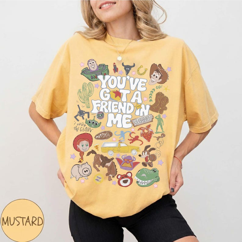 Op de afbeelding: Een mosterdgeel T-shirt met korte mouwen en een grafische print met de tekst "YOU'VE GOT A FRIEND IN ME", omringd door diverse ge&iuml;llustreerde personages en iconen uit de Toy Story-franchise, waaronder Woody, Buzz Lightyear en Jessie.