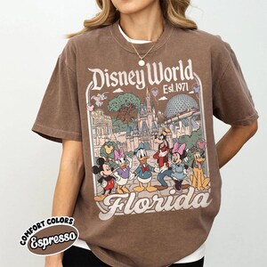 May include: Brown t-shirt featuring a Disney World graphic with Mickey Mouse, Donald Duck, and other characters. The design includes the text "Disney World Est. 1971 Florida". The shirt is labeled "Comfort Colors Espresso".