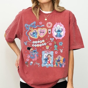 May include: A maroon t-shirt featuring a colorful graphic design with the text "LOLO STITCH". The design includes cartoon characters in frames, bows, and floral elements. The shirt is made of a soft material.