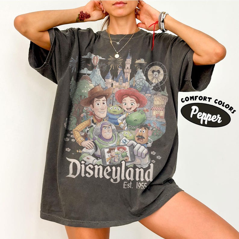May include: A charcoal gray short-sleeve t-shirt featuring a graphic print of Toy Story characters including Woody, Jessie, Buzz Lightyear, Rex, and Mr. Potato Head in front of a Disneyland castle. The text reads Disneyland Est. 1955 and Comfort Colors Pepper.