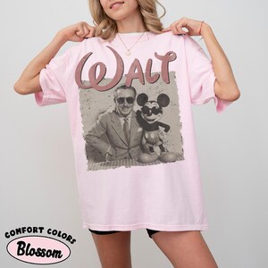 May include: Light pink t-shirt featuring a vintage-style graphic of Walt Disney with Mickey Mouse. The word "Walt" is arched across the top in a matching pink hue. The shirt is labeled "Comfort Colors Blossom".