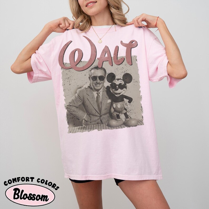 May include: Light pink t-shirt featuring a vintage-style graphic of Walt Disney with Mickey Mouse. The word "Walt" is arched across the top in a matching pink hue. The shirt is labeled "Comfort Colors Blossom".