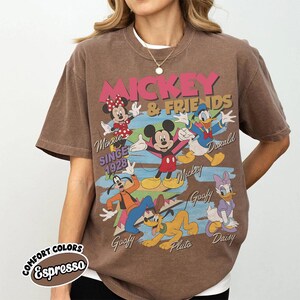 Peut inclure: T-shirt marron avec un motif de Mickey Mouse et ses amis, dont Minnie, Donald, Dingo, Pluto et Daisy. Le design comprend le texte "Mickey & Friends" et "Since 1928". Le t-shirt est de couleur "Espresso".