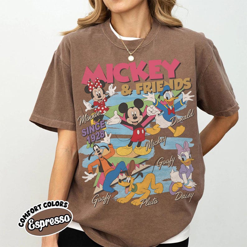 Peut inclure: T-shirt marron avec un motif de Mickey Mouse et ses amis, dont Minnie, Donald, Dingo, Pluto et Daisy. Le design comprend le texte "Mickey & Friends" et "Since 1928". Le t-shirt est de couleur "Espresso".