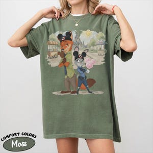 Puede incluir: Camiseta Comfort Colors verde musgo con una ilustraci&oacute;n de dibujos animados de Nick Wilde y Judy Hopps de Zootopia, con orejas de Mickey Mouse. La imagen incluye un castillo, un sol y el texto "Comfort Colors Moss".