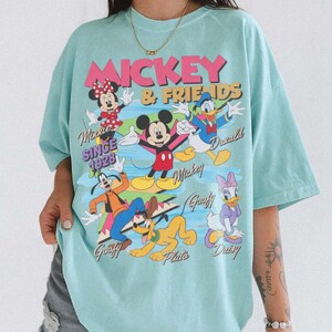 Peut inclure: T-shirt bleu clair avec un motif de Mickey et ses amis. Le graphisme comprend Minnie, Mickey, Donald, Dingo, Pluto et Daisy. Le texte indique "Mickey & Friends" et "Since 1928".